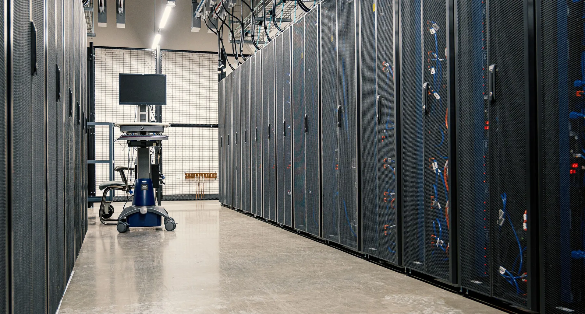 An aisle between two rows of server cabinets in a data centre, cabling visible through the mesh doors.