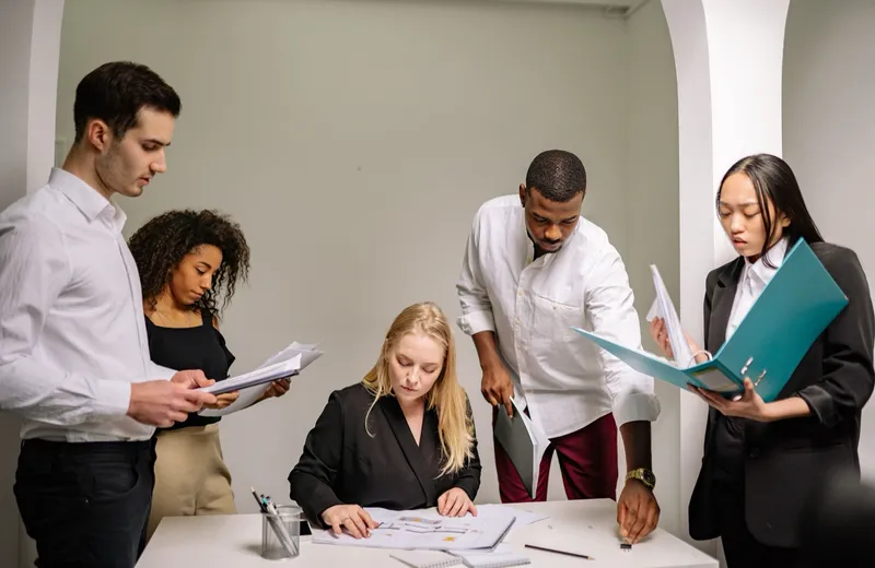Five colleagues around a desk, reading through printed documents and folders together.