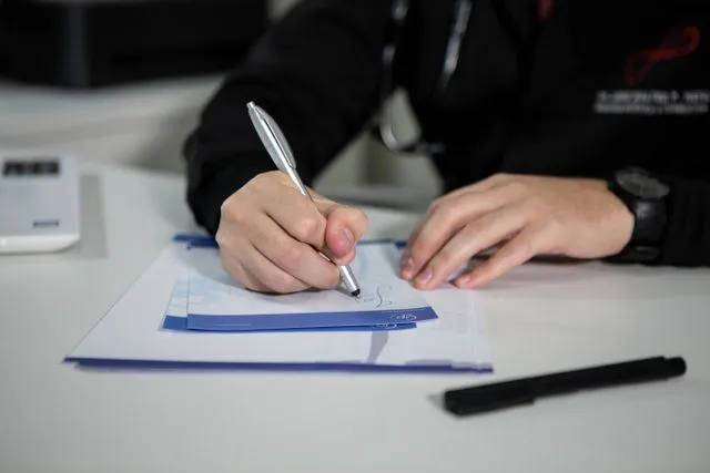 A biller at a desk completing a claim form by hand, pen in hand over a stack of forms.