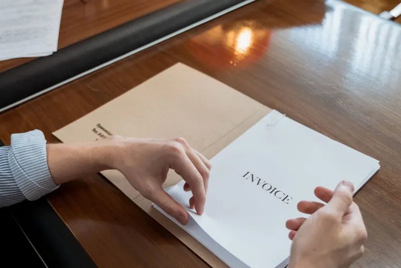 Hands easing a document headed Invoice out of an envelope on a wooden desk.