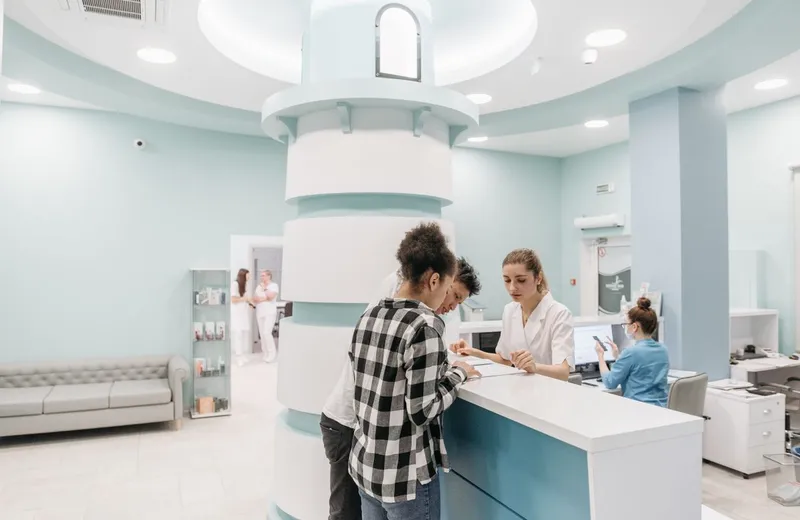 A clinic reception desk, a patient checking in while staff work at the counter behind.