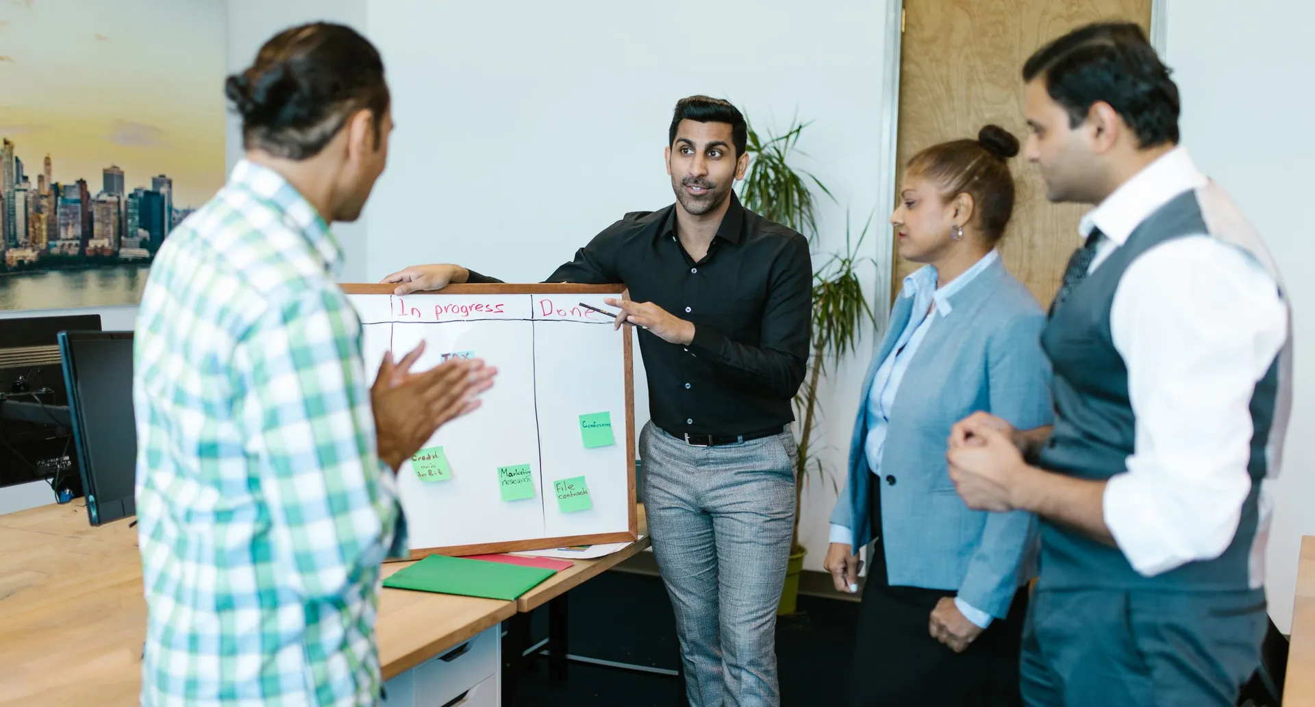 Four colleagues standing at a whiteboard divided into progress columns, covered in sticky notes.