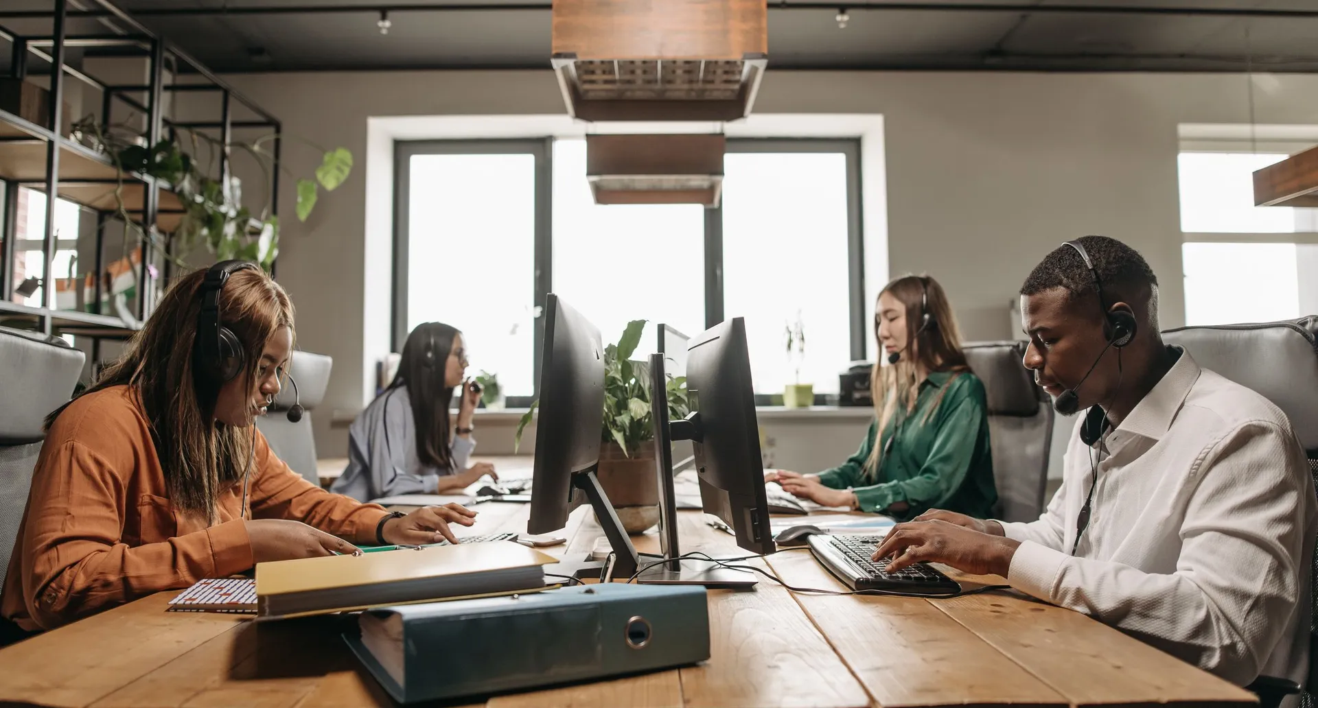 Four people working at one long shared desk with monitors and telephone headsets, tall windows behind them.