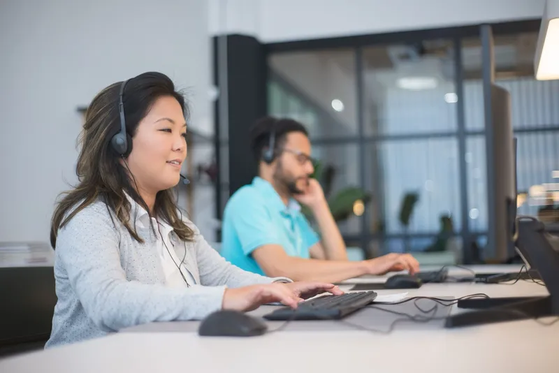 Two people working at desktop monitors, both wearing telephone headsets.